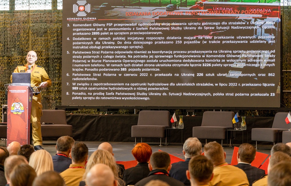 Strażak stoi na scenie, przed mównicą. Przemawia do zgromadzonej publiczności.
