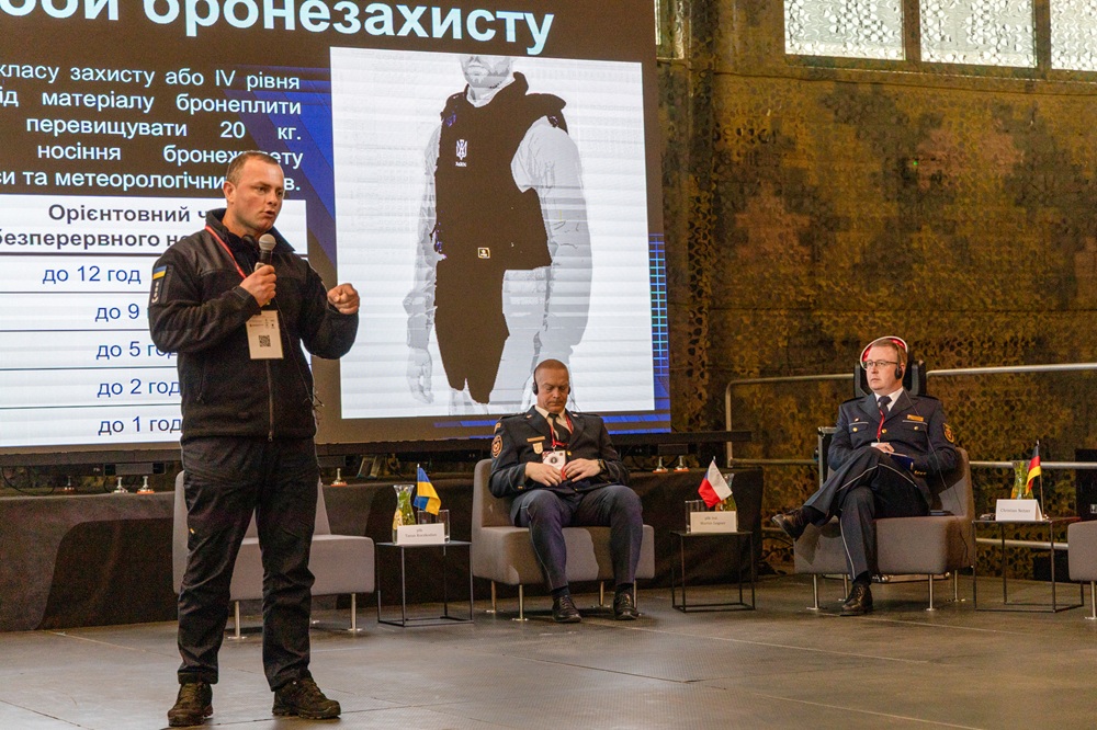 Mężczyzna stoi na scenie. Mówi do mikrofonu. Na scenie siedzą również dwaj mężczyźni.