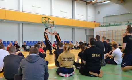 Sala gimnastyczna. Dwóch mężczyzn prowadzi ćwiczenia. Reszta osób siedzi bądź klęczy na podłodze.