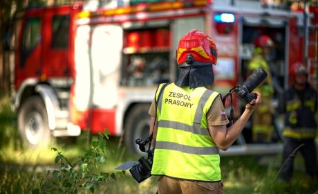 Strażak w jaskrawej kamizelce z napisem "Zesp&oacute;ł prasowy" stoi z parartem w ręku.