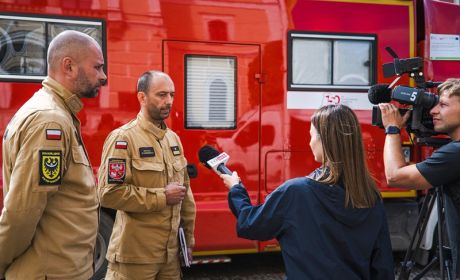 Dziennikarka prowadzi wywiad z dwoma strażakami. Obok dziennikarki stoi oparator i nagrywa rozmowę.