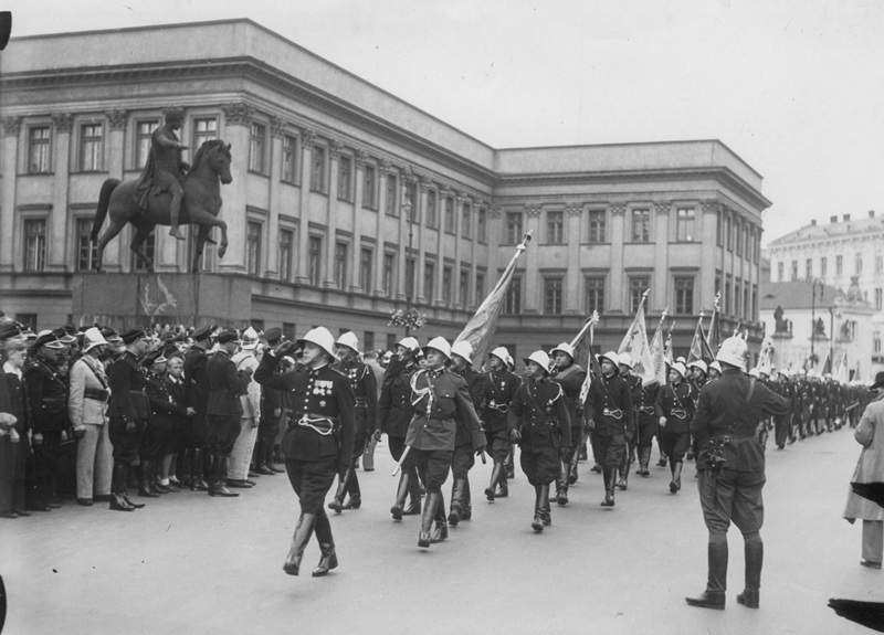 Czarno-białe stare zdjęcie. Delagacja strażak&oacute;w maszeruje w kierunku Grobu Nieznanego Żołnierza. Kilku strażak&oacute;w niesie sztandary. W oddali widać budynek i pomnik.