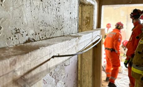 Na ścianie znajduje się belka. W oddali widać kilku strażaków w pomarańczowych ubraniach.